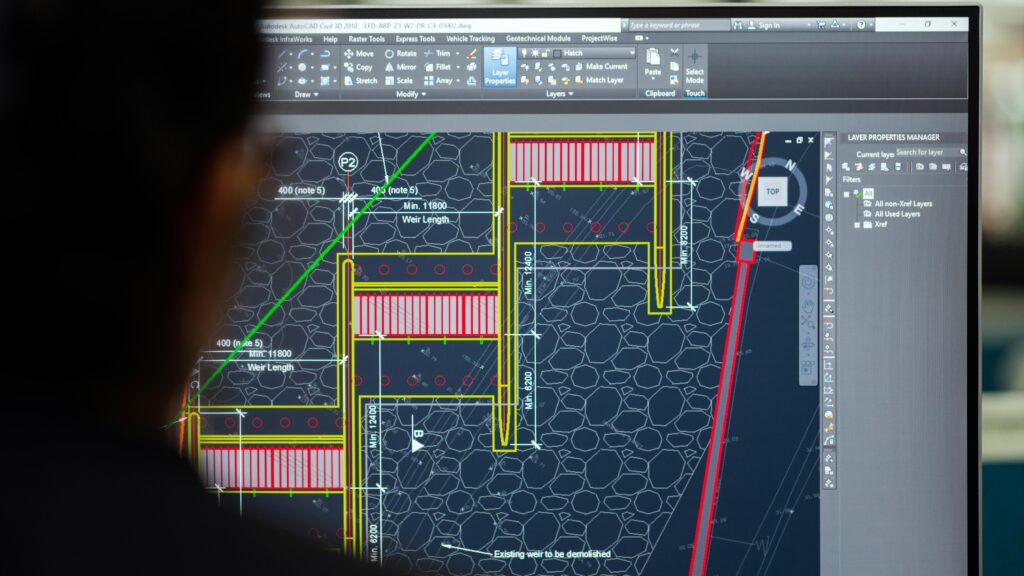 A civil engineer working on a weir design using CAD software on a computer screen in an office setting.